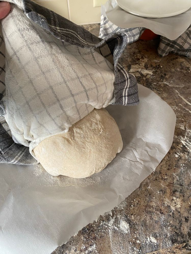 A round loaf of dough being turned out on a piece of parchment paper on the counter. A flour dusted cloth is being peeled off of the dough. 