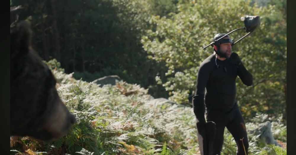 A forest scene with a fake bear head in the left of frame and Allan Henry standing on the right in black Lycra, a head rig with silicone snout and arm extensions 