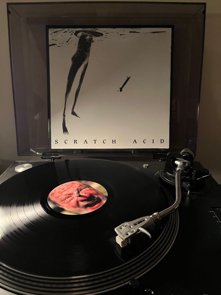 Scratch Acid’s S/T and Berserker EP playing on my turntable. The vinyl platter is black with a close up of lead singer David Yow’s face. The album cover is white, with a black charcoal sketch of a skinny, humanoid female from the shoulders down on the left hand side.