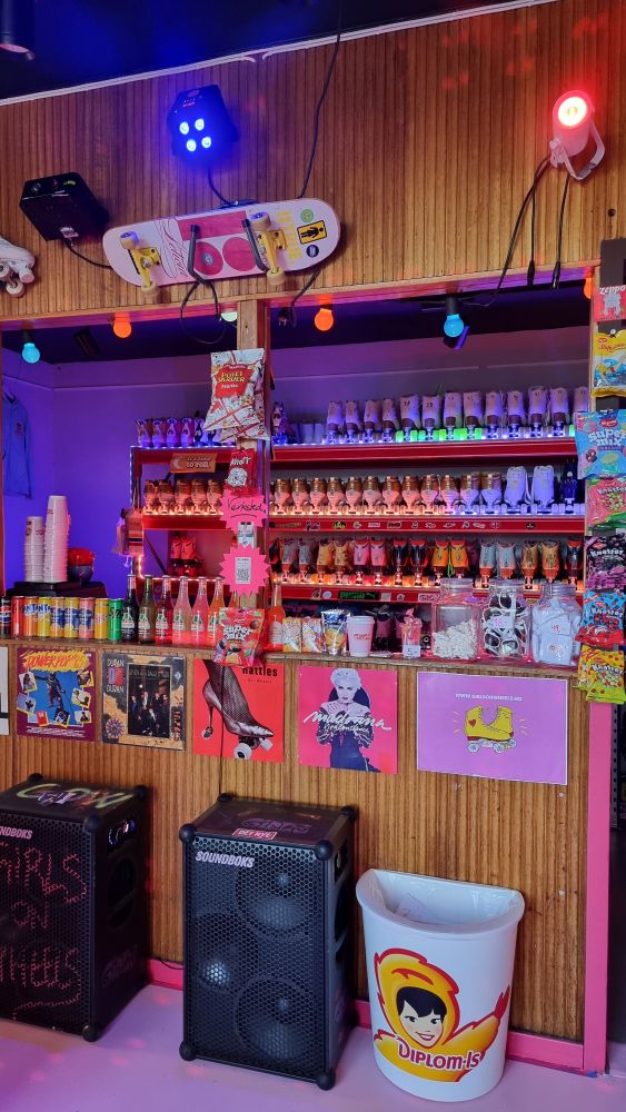 Small cafe with sodas, candy, chips, sunglasses in a jar. Posters in many colors, rollerskates in the shelves in the back, big speakers in the front. Disco lights on the wall.