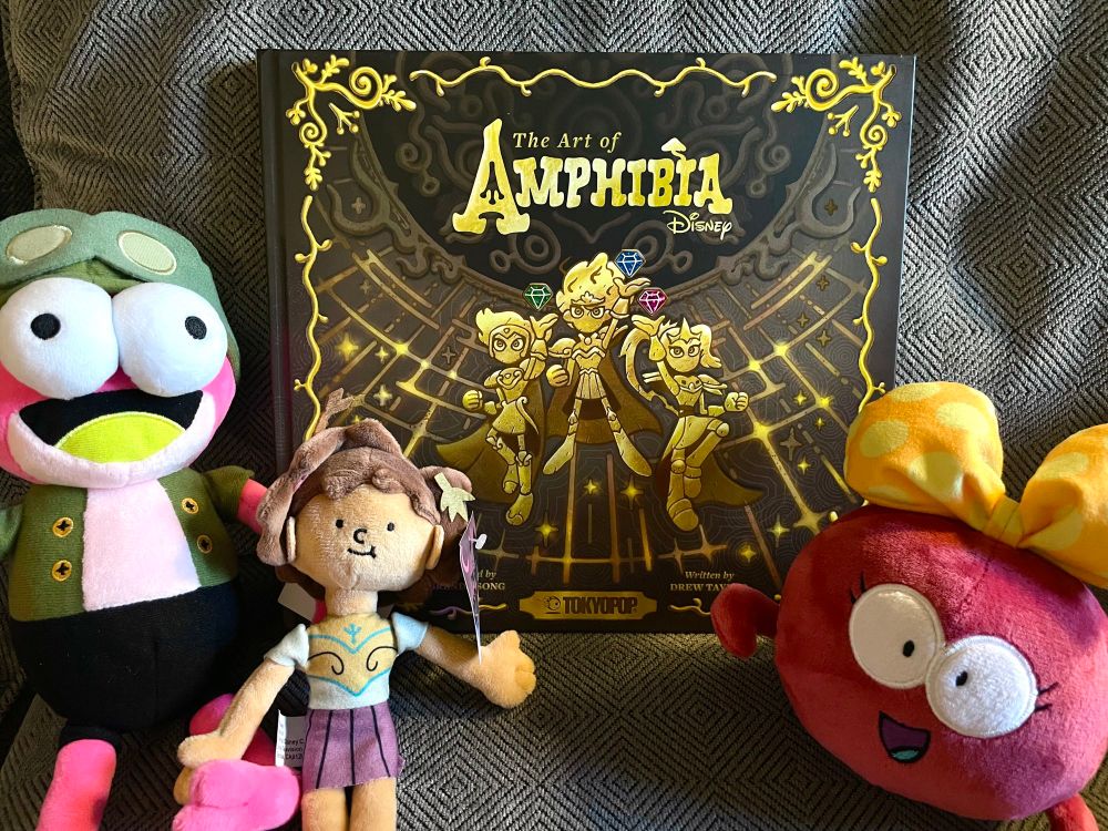 A picture of the Art of Amphibia book surrounded by plushies of characters Sprig, Anne, and Polly.