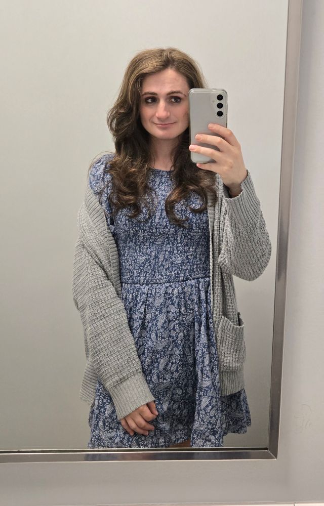 Ashes in front of a mirror at work, she is wearing a blue dress with a white patten and her grey oversized cardigan