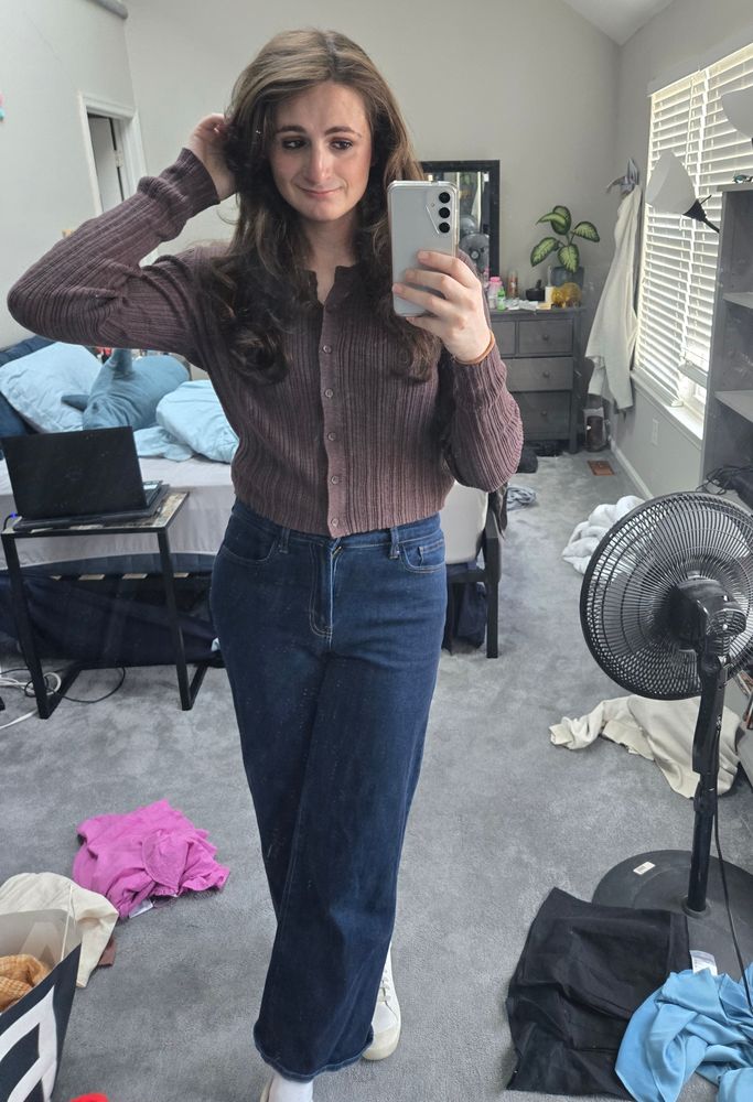 Ashes in front of her full length mirror at home. She is wearing dark blue jeans and a brown lightweight sweater/shirt. Her hand is brushing a piece of hair back behind her left ear