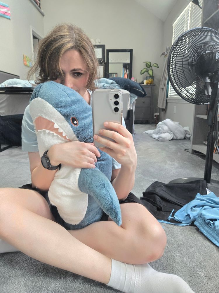 Ashes sitting on the floor with her legs crossed and holding a stuffed shark (blahaj)