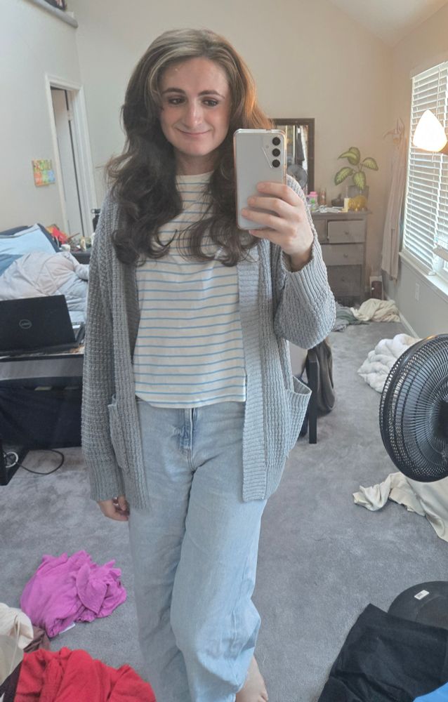 Ashes in front of her full length mirror. She is wearing light blue jeans, a white shirt with blue stripes and an overiszed grey cardigan