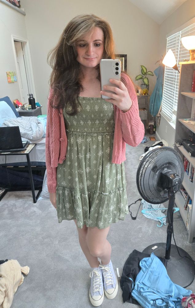Ashes in a green dress with a white pattern on it, pink sweater unbuttoned, and blue converse. Her room has lots of stray articles of clothing just hanging about.