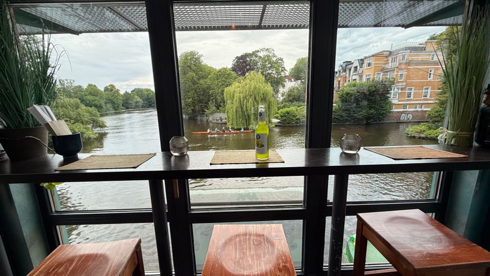 Barstools set along a counter with floor to ceiling glass windows giving a lovely view over the waterways of Hamburg. 