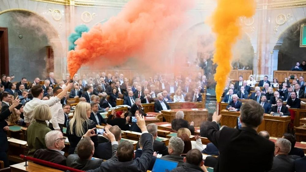 A parliamentary session is disrupted as protesters inside the chamber release orange and green smoke from handheld smoke flares. Lawmakers, dressed in formal attire, react with surprise, some taking photos or recording the event. The ornate chamber features wooden desks and intricate architectural details. The atmosphere appears chaotic, with attention focused on the protesters holding the smoke flares.

