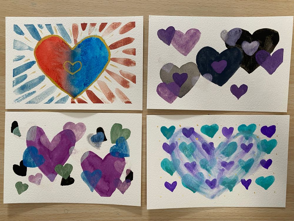 4 water color paintings on a board. Upper left: large heart with red (left) and blue (right) halves with a melded center. Streaks of blue, red and gold surround it. Upper right: a trio of larger hearts (left to right grey, dark grey, black) anchor an arrangement of Purple Hearts of various sizes and inks, including an almost invisible purple pearlescent. Bottom left: 2 large Purple Hearts anchor an arrangement of black, green, purple and pink smaller hearts. Bottom right: a grid of small blue and purple alternating hearts with gold dots between them has a larger heart painted on top with pearlescent paint. This paint picked up the colors of the hearts as it was painted . 