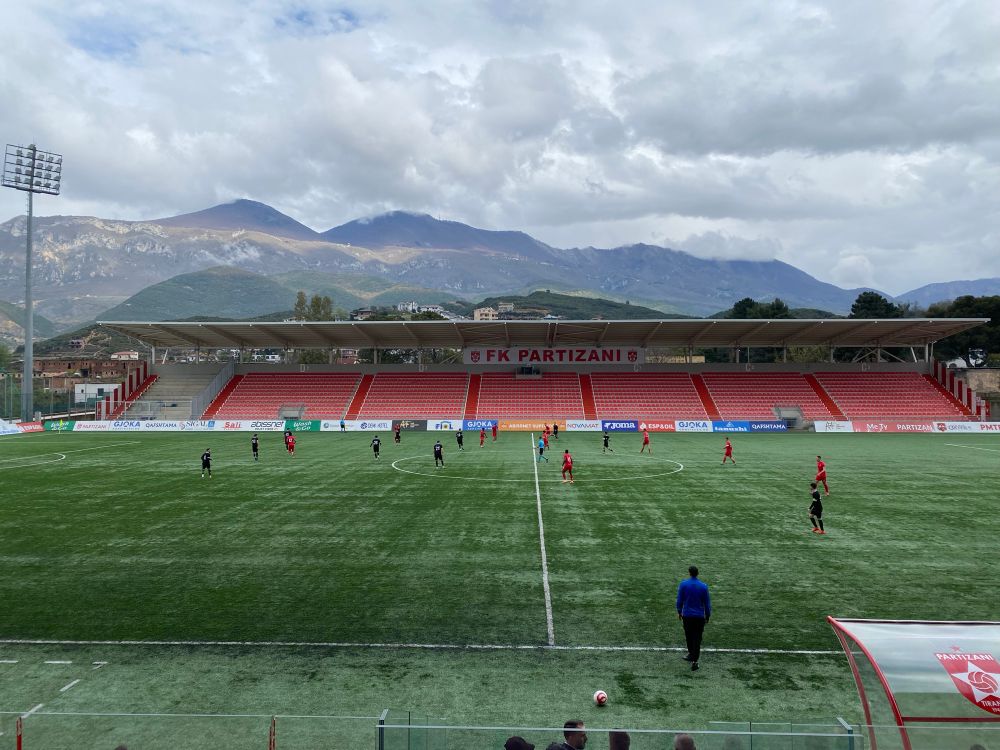 Stadion Partizani in Tirana, die Tribüne, ein Flutlichtmast, und ein Gebirge sind im Hintergrund zu sehen, im Vordergrund sieht man die Spieler und das Spielfeld 