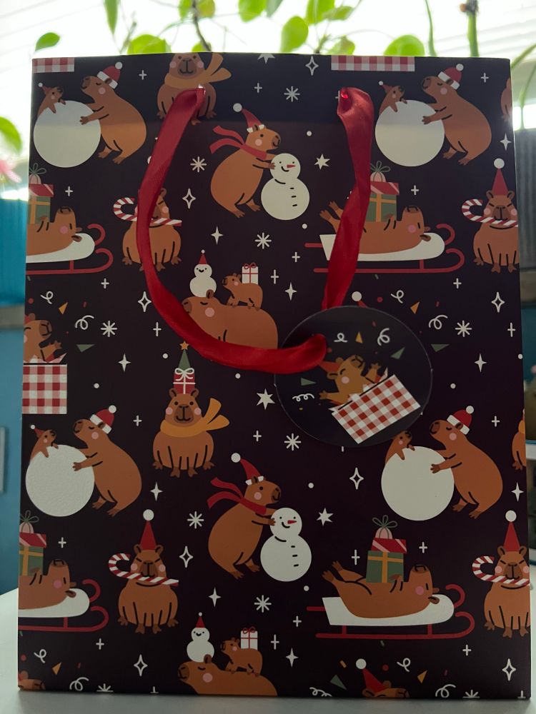 Eine weihnachtliche Geschenktüte auf der viele Capybaras in verschiedenen Weihnachtssituationen abgebildet sind. Die weißen Schneeflächen glitzern 