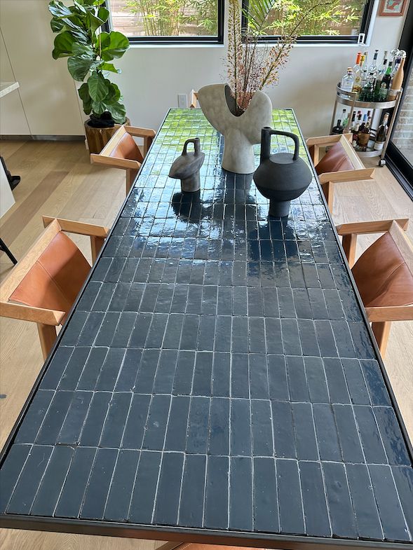 Custom rectangle dining table by MK Workshop with a dark navy blue zellige tiled surface, bordered by a slim blackened steel frame. The glossy rectangular tiles are neatly aligned in a grid pattern, reflecting light subtly. Surrounding the table are caramel-colored leather chairs with light wood frames.