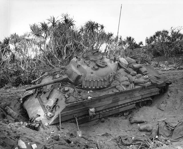 Black & white picture of an M4A3 Sherman tank stranded in a sandy shell hole with bushes in the background. The sides of the hull are covered in wooden boards. There's a thinner wooden board covering the length of the suspension too. The top of the tank hull is covered with sandbags.

The turret has a length of spare track attached to its side, while the turret front, gun mantlet and half of the gun is covered in a dark material. This is possibly a light canvas covered in a waterproofing agent. Traces of this dark material are also visible around the turret pistol port on the turret left-hand side and around the hull MG port. The tank, including the wooden boards and spare track, has been painted in a two-tone camouflage scheme.
