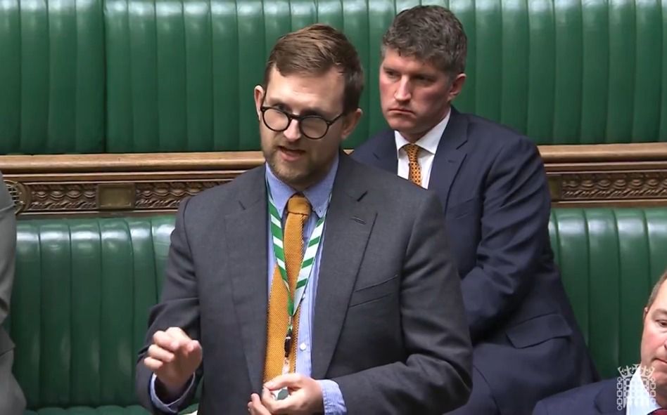 White man in grey suit and yellow tie makes speech in parliament.there is another man sitting on the green bench behind him. 