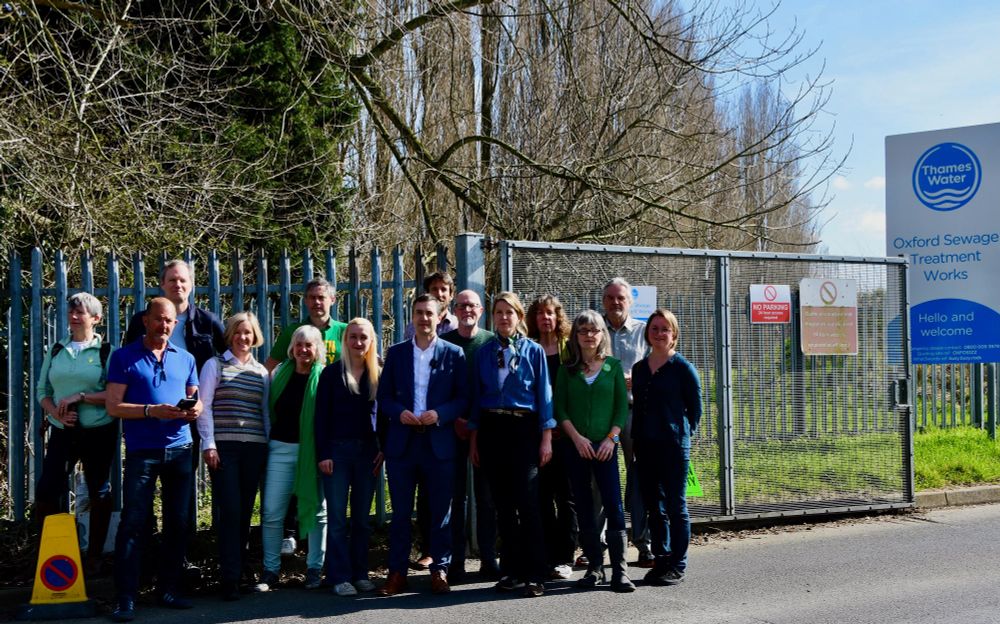 People, many of them wearing green, standing outside the gate to Thames Water Sewage Treatment Works . Adrian Ramsey, in a suit, not wearing green, stands in the centre. (via Green Party)