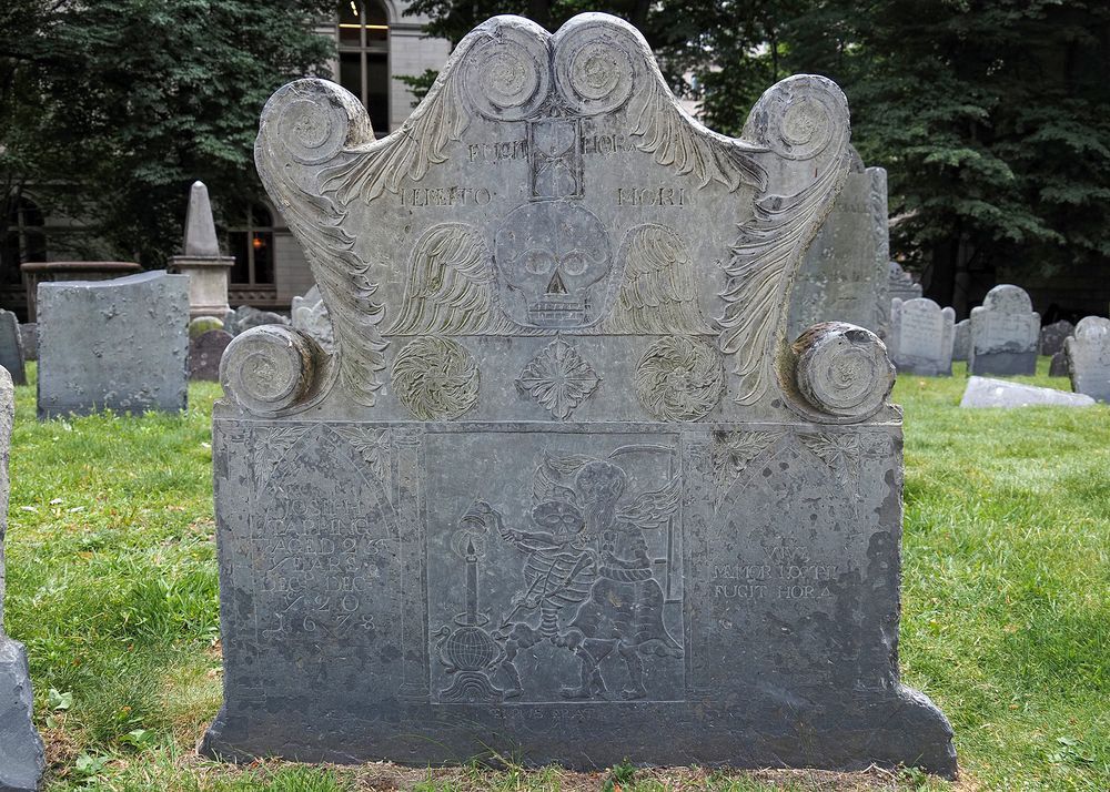 An elaborate slate headstone featuring multiple motifs representing "memento mori", including a winged skull with hourglass on top, and beneath a depiction of a struggle between a skeleton and Father Time. The skeleton is armed with an arrow and a candle snuffer, and is attempting to put out a large candle. 
