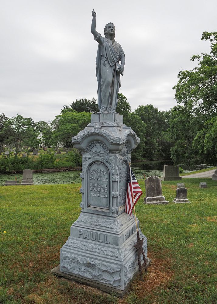 A white bronze monument featuring a pedestal with classically dressed figure standing on the top. One arm is reaching towards the sky. 