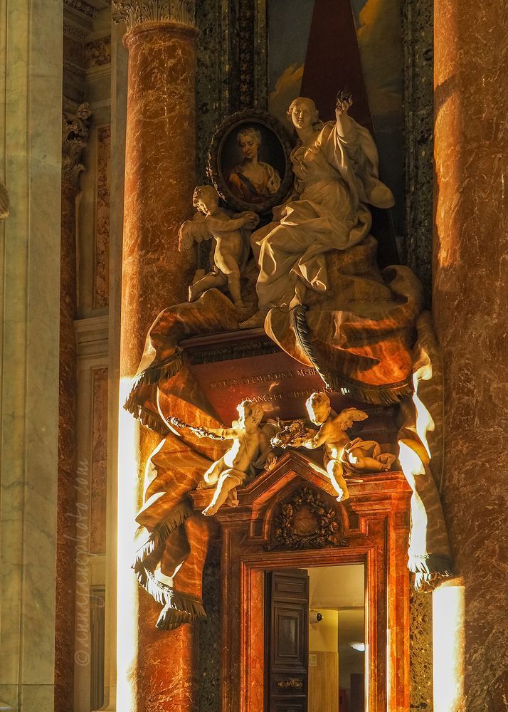 Another angle of the monument shows additional cherubs, and that the monument is situated above a doorway. The photo has a orange tint due to the sunrise.