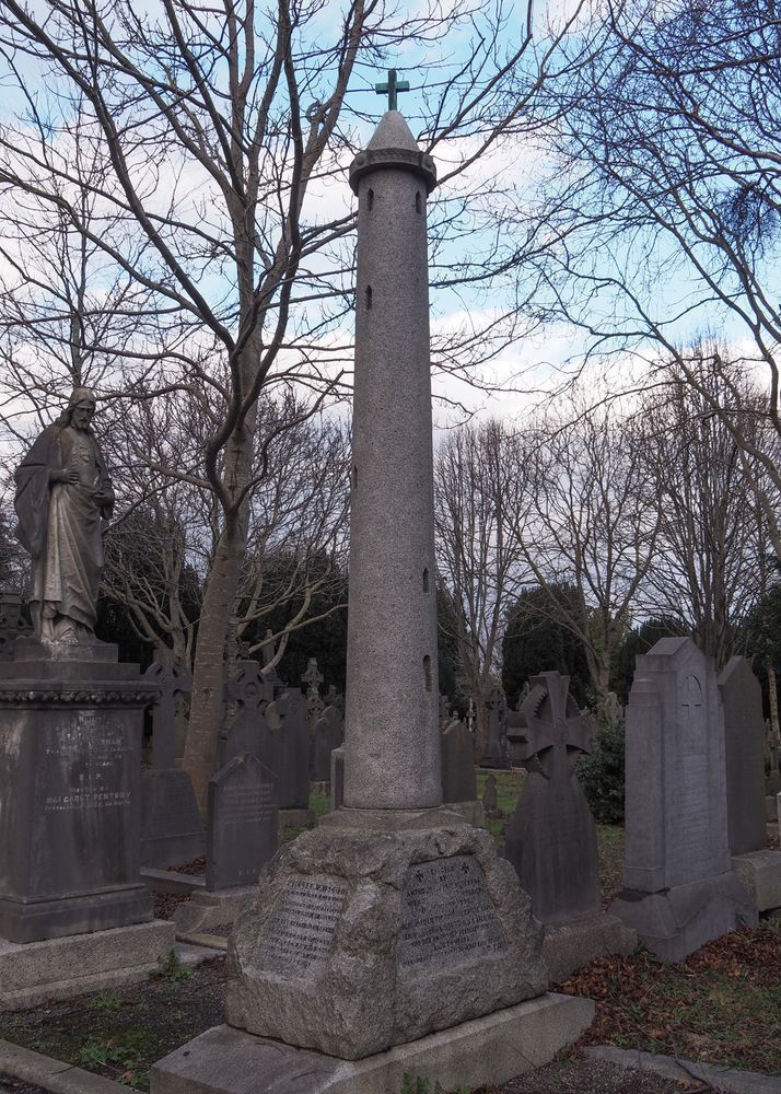 A granite cemetery monument in the shape of a round tower, topped with a cross. It is mounted on a rock shaped base and is surrounded by other memorials.