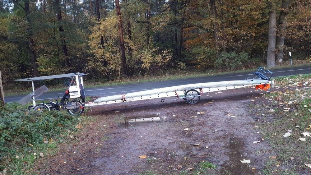 Ein Lastenrad mit einem Fahrradanhänger, auf dem eine Leiter befestigt ist, steht an einer Straße.