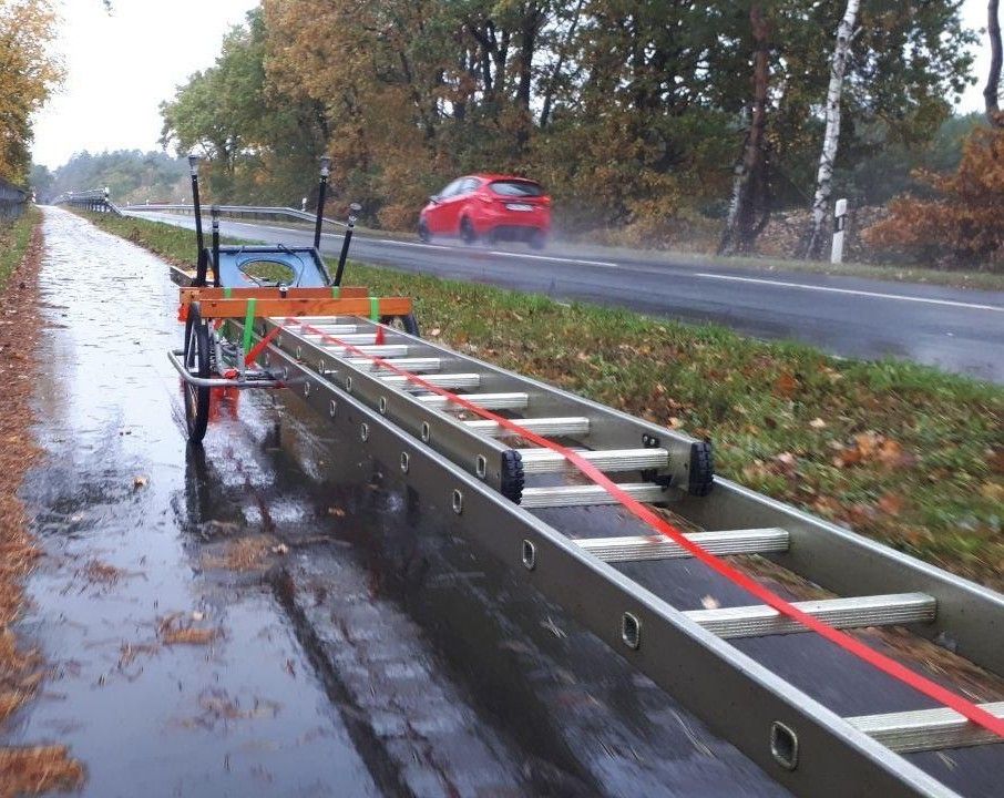 Ein Fahrradanhänger steht an einer regennassen Straße. Darauf befestigt ist eine lange, ausgezogene Leiter.