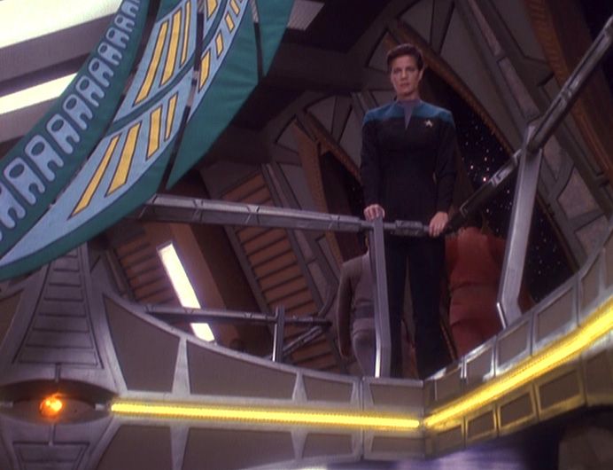 Screenshot from Star Trek Deep Space 9, season 4 episode 6 "Rejoined".  Dax is standing on the upper level of the promenade looking forlorn.  A banner is draped, hanging from something offscreen (the ceiling?) to the side of the railing next to Dax.  The pattern on the banner is irrelevant to the story, but along the banner's edge is a series of repeated shapes that are rather reminiscent of Among Us crewmates.