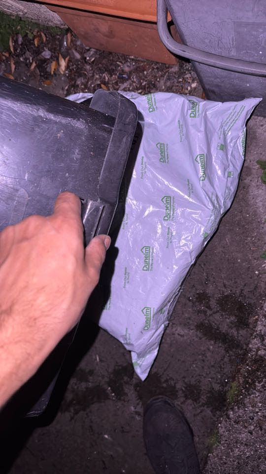 A plastic-wrapped parcel from Dunelm. Someone (the courier) is upending one of my recycling bins to put over it.