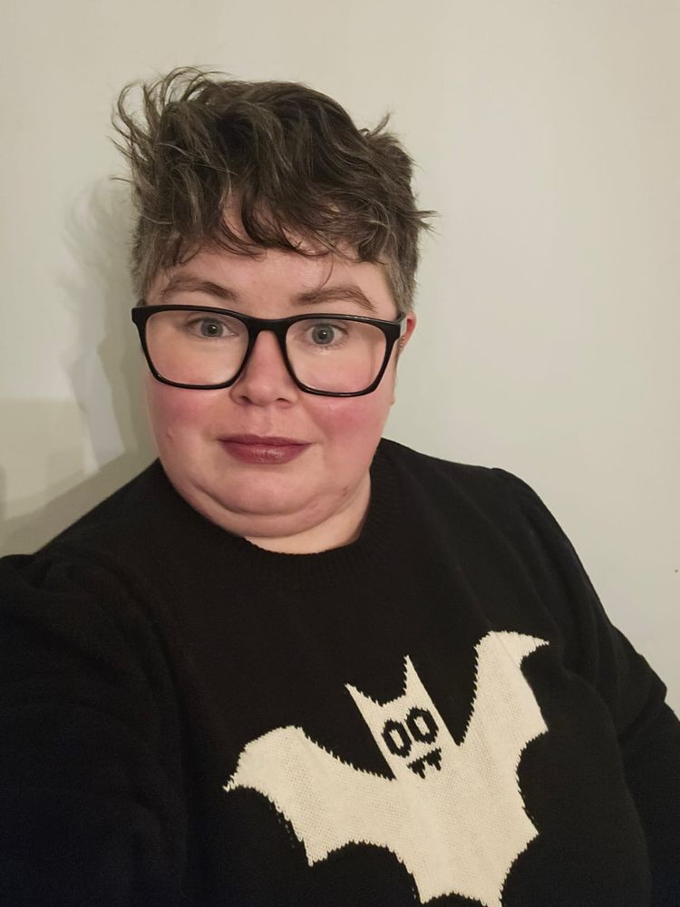 Sandra, a white femme person with short wavy brown hair and black glasses, smiling at the camera. They are slightly flushed from the cold outside. They are wearing purple lipstick and a black jumper with a white cartoon vampire bat on it.