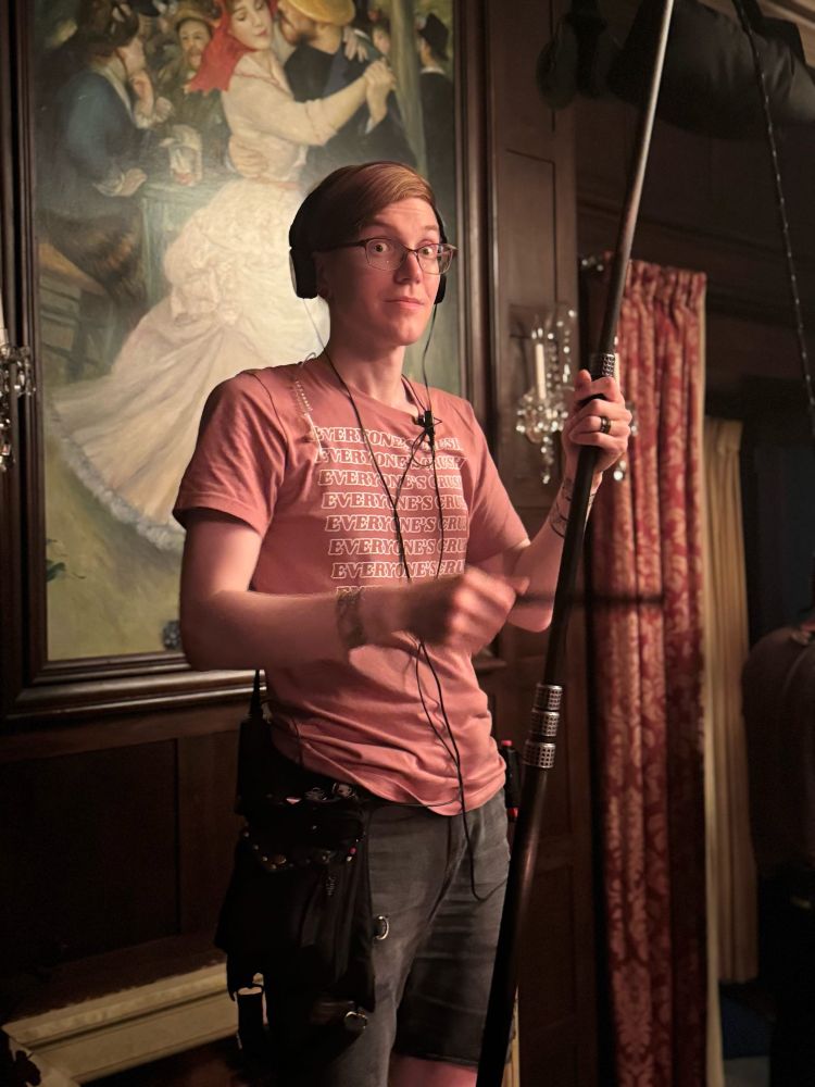 Cait looks surprised at the photo being taken. She holds an extendable stick next to her that goes out of frame. Cait wears black shorts, a black belt pouch, and a pink shirt that repeatedly says “everyone’s crush”.