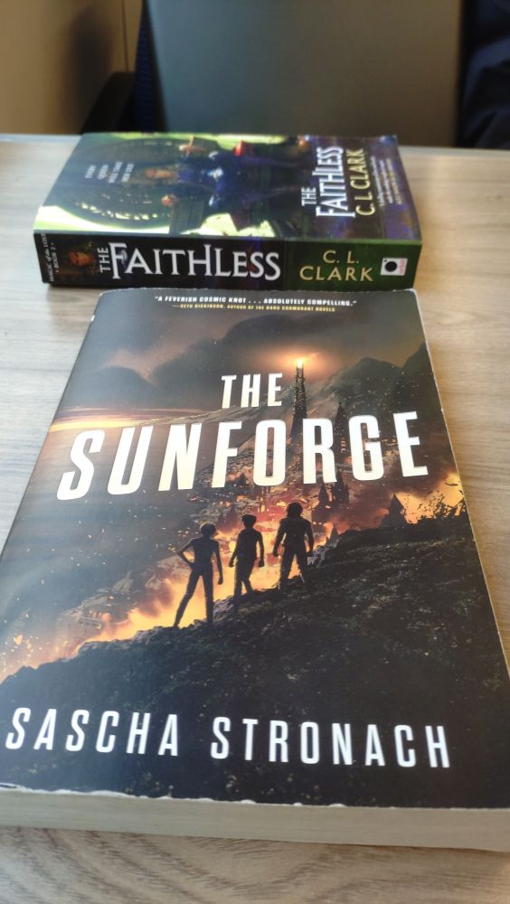 Book cover on train carriage table, The Sunforge by Sascha Stronach

Art shows three people silhouetted on a hill looking down over a burning coastal city with a tower in the background with flames on the top-vaguely similar to Sauron's tower which I'm sure isn't a coincidence

Behind is another book side on, The Faithless by C.L. Clark, starting that now 