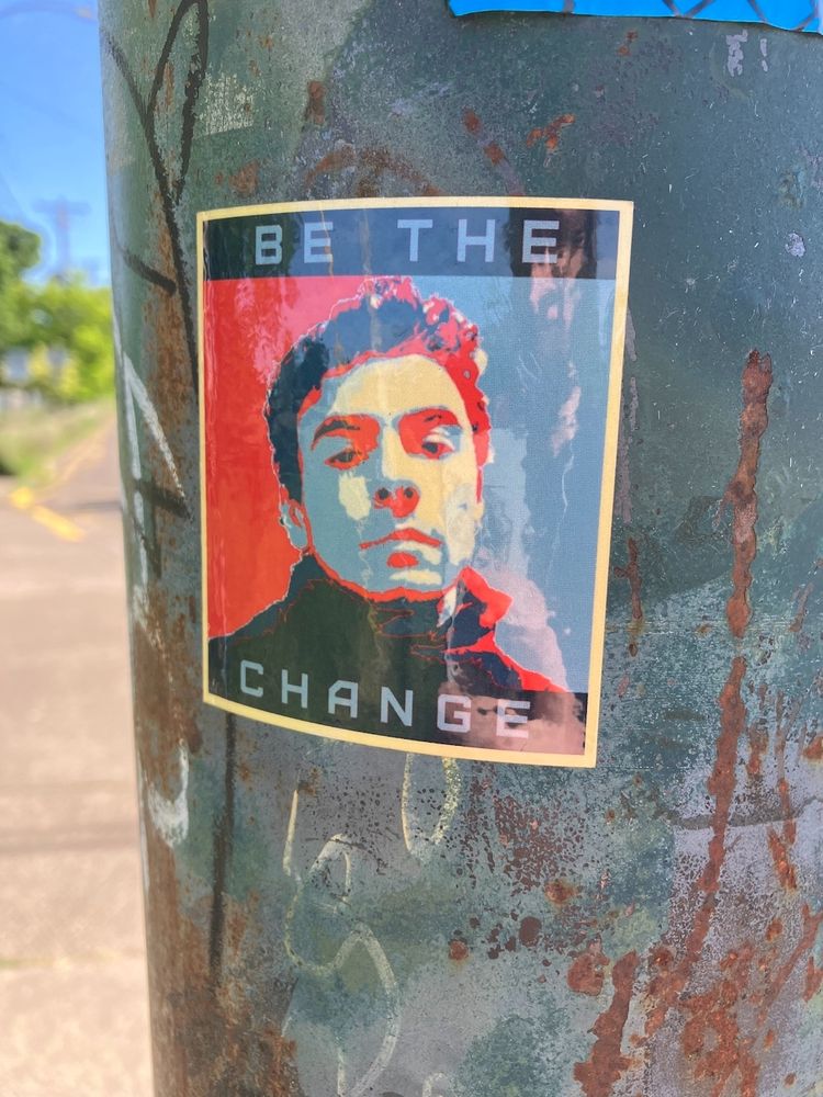 Photo of a sticker on a pole. Sticker is an illustration of Luigi Mangione with the words "BE THE CHANGE" 