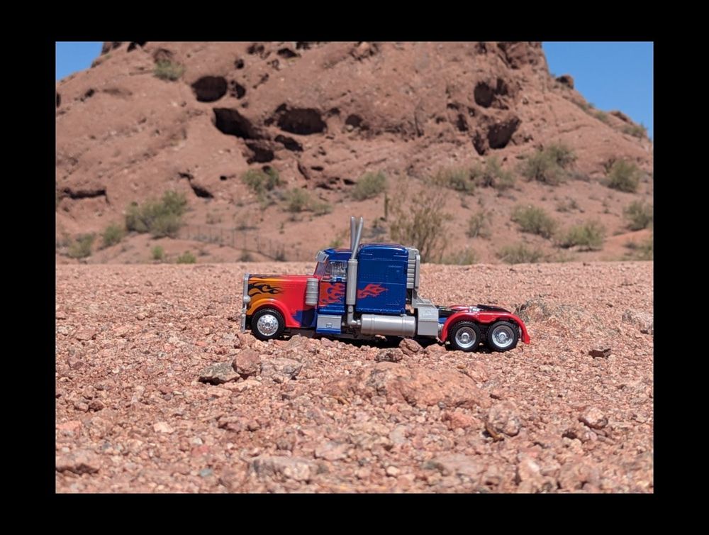 A blue and red truck with painted flames on the side. The background is a desert with a small mountain in the background 