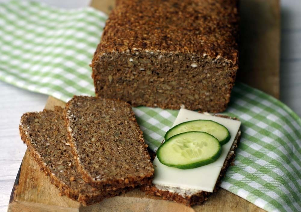 Ein angeschnittenes Schwarzbrot auf einem Brett. Unter dem Brot ist eine grün weiß karierte Serviette gefaltet. Es sind drei Scheiben abgeschnitten. Zwei davon liegen aufgefächert nebeneinander. Daneben eine einzelne Scheibe, die mit Käse und Gurken belegt ist.