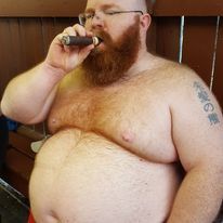 A bald man with a red beard and glasses sits shirtless against a fence in a bar. He is smoking a cigar.