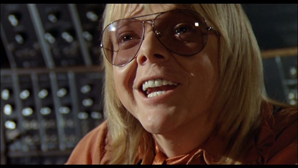 A man in front of a sound board.  He had longish blond hair and a toothy smile.  He is a Caucasian wearing light tinted sunglasses and an orange shirt