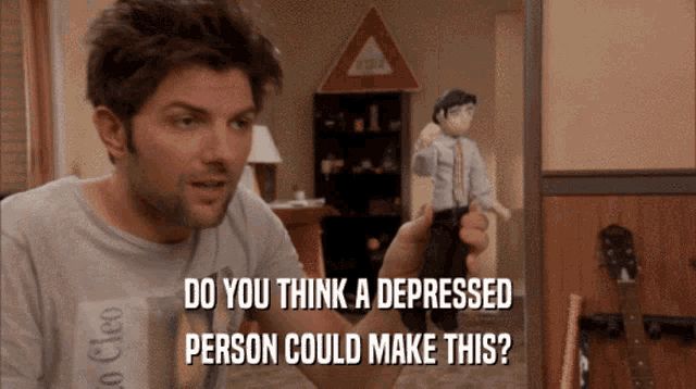 Adam Scott as Ben Wyatt in Parks and Rec , looking dishevelled, holds out a handmade doll of himself, subtitles show him saying “do you think a depressed person could make this?”