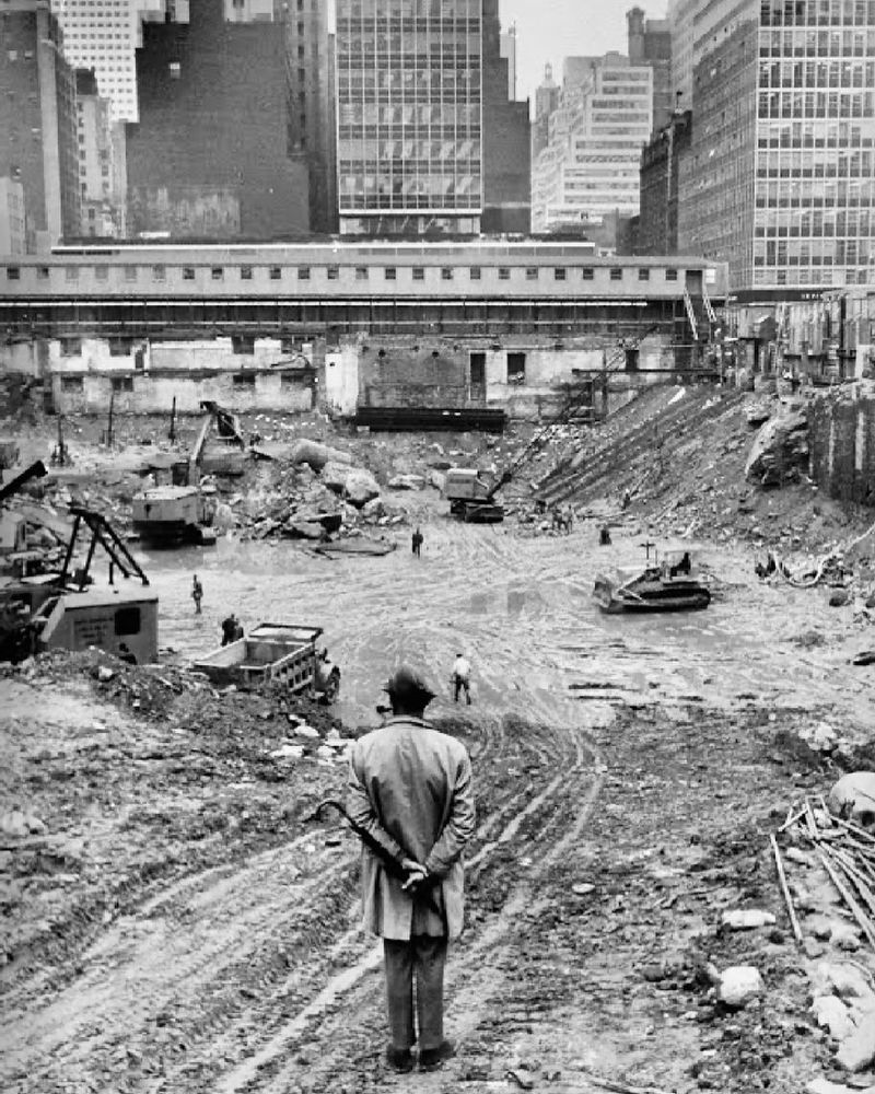 📷 Yale Joel 
We see the back of Jacques Tati as he looks down on a big wide hole being dug out in preparation for a building in a city. We see diggers, trucks, bulldozers and workmen in the hole. Further on at the top of the photograph we see skyscrapers blocking most of the sky. He stands bottom centre, legs close together, coat, trousers, smart shoes, hands behind his back, pipe sticking out from his mouth to his left just so we can see it, trilby hat.