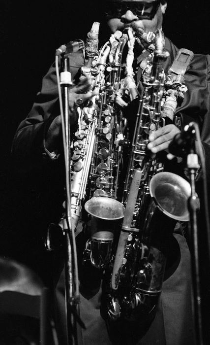 📷 Max Oettli 
A black & white close up photograph of Roland Kirk playing multiple instruments at the same time. Looks like three saxophones. We see from the top of his black glasses to the bottom of his Saxophones. The rest is black.