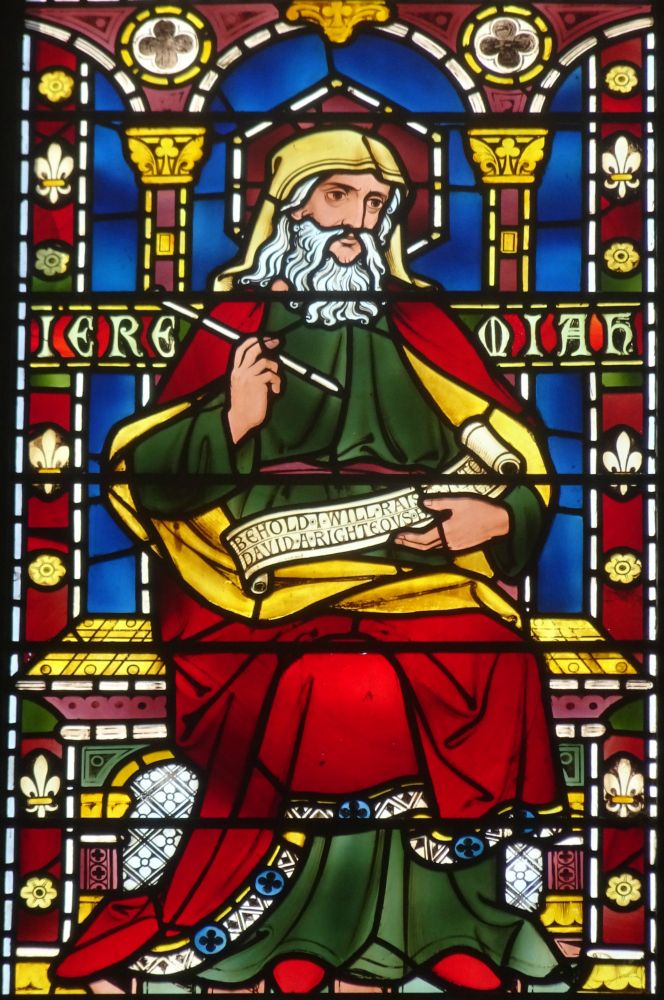 Stained glass window by Alexander Gibbs of Jeremiah about to write something down on his scroll. He is paused as if thinking about what to write.
