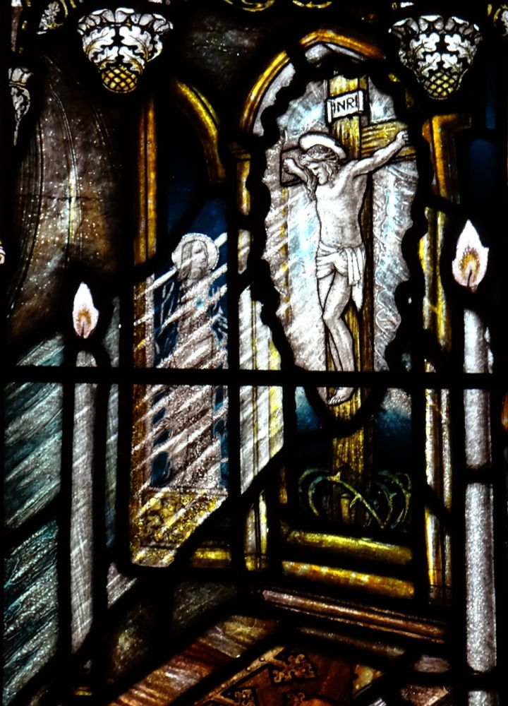 Detail from the stained glass window in the quoted post showing two parts of a tryptych painting. Jesus on the cross in the centre with Mary looking up to him on his right. Light is shown to emanate from him in streams almost concealing the figure of Mary. In front of the painting is a lectern with a bible on it and on either side are two long lit candles.