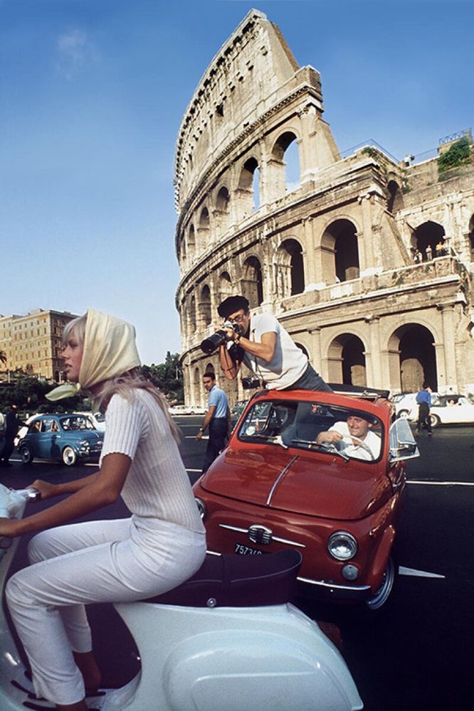 📷 Douglas Kirkland 
Colour photograph of Brit Ekland in white on a white Vesper being chased by a photographer (Peter Sellers) standing through the open top of a red car trying to get his picture as the driver swings the car round a tight corner on two wheels, it looks almost. Behind is the Colosseum. However it was done a lot of trouble has gone into the joke.