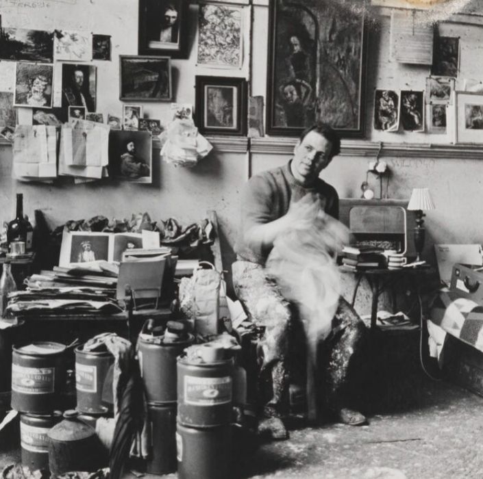 📷 Snowdon 
Black & white photograph of Frank Auerbach in his cluttered studio. It includes a familiar feature from some of Snowdon's theatre photographs, the blur. Here Auerbach is sitting wiping his hands on a cloth vigorously such that, arms, hands and cloth are a blur. He looks up, to his right with his head leaning to his left, he could be looking out of a high window, or simply musing. The blur is a feature of Auerbach's work too. Behind him, pictures are attached to the wall, some are his works but I also recognise a print of Titian's blue man and I think a tiny Rembrandt self-portrait as Zeuxis laughing, which would make sense as it is Rembrandt doing an Auerbach.