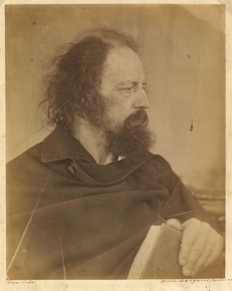 📷 Julia Margaret Cameron
Albumen print. Brown tones. Almost fully a profile shot, we see a glimpse of Tennyson's left eye as he looks to his left and down. He holds a book in his left hand in his lap but towards us so the book and hand occupy the right hand corner and are closest to the camera. He wears a cloak the collar of which curves round his neck and on to his left shoulder. With this pose we can now see the back of his dark frizzy hair, wind blown in appearance, against the lighter coffee cream colour of the smooth background wall or screen.