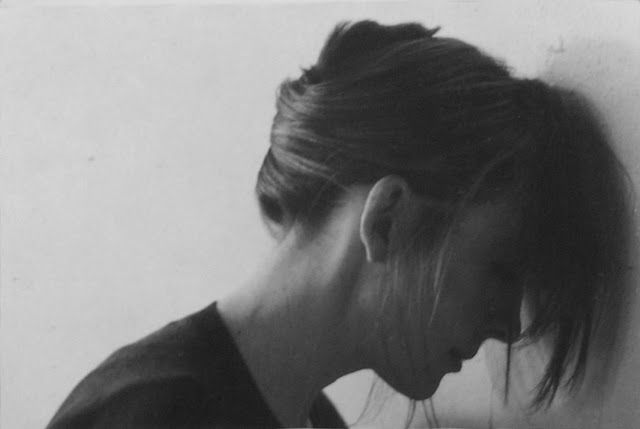 The third of three photographs in this thread of Diane Keaton by Annie Leibovitz. Black & white. With her hair tied up at the back but long and loose at the front we see Keaton in profile leaning her head and hair against a white wall and casting on it a curved light grey shadow, with the side of her face a darker shadow. All else is just her neck and shoulder in a black top extended behind her and curving down beyond the base of the photograph.