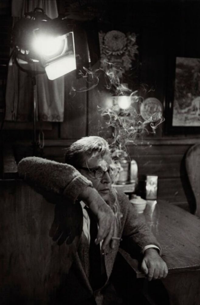 📷 Bob Willoughby 
A black & white photograph taken during filming of Who's Afraid of Virginia Woolf. We see Richard Burton side-on with his arm high on the back of a chair, cigarette between his fingers, smoke spiralling up to form a tree-like crown of interlocking smoke trails just above his head lit by a spotlight on the top left corner, his other arm resting on a table, his fingers holding the edge. As he looks to our right he could be enjoying a break, mentally rehearsing his lines, or acting in front of a camera rolling.