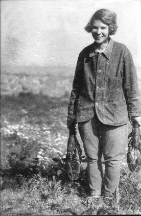 📷 unknown to me 

A black & white photograph of Margaret Fay Shaw standing in moorland, her feet disappearing in the ground, her boots or glasses in her hands, she stands to the right of the photograph, her left arm clipped by the right edge, on the left, numerous small white flowers, then the distant flat moorland, and above the light grey sky. Her body ever so slightly turned to our left, she looks at the camera grinning.