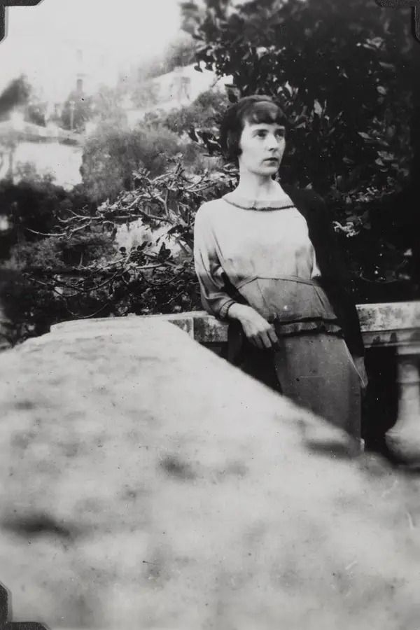 📷 Ida Baker
Black & white photograph of Katherine Mansfield leaning back against a balustrade looking up to our right, but much of the photograph is taken up with the top of the balustrade going away from us as if the camera was resting on it as the picture was taken. Then in the middle of the picture the balustrade turns right and we find Mansfield there in the corner as it curves round. Behind her are trees on the right and a view of hills then sky on the left. 