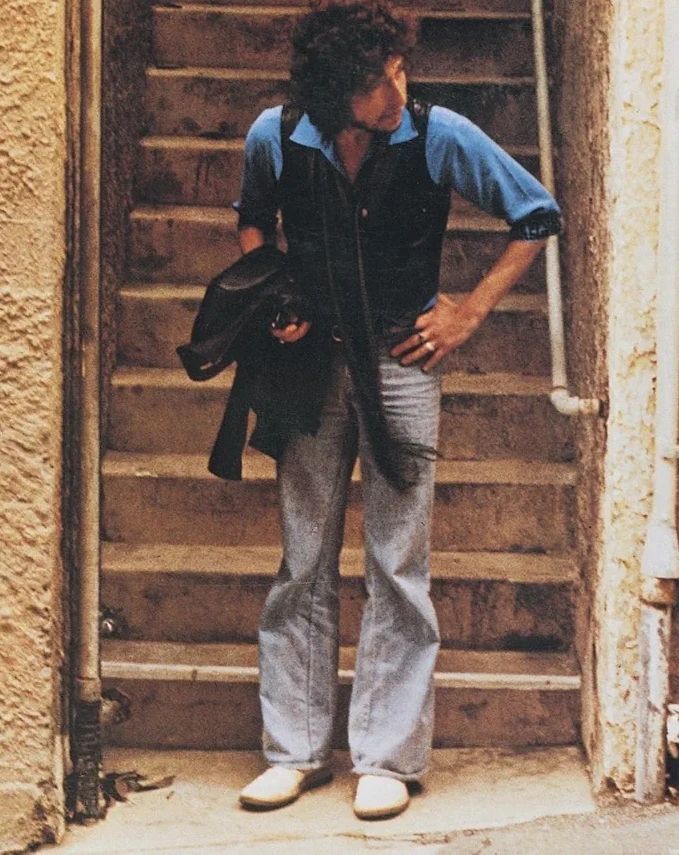 📷 Howard Alk
A cropped colour photograph of Bob Dylan at the bottom of some stairs poking his head past a wall to look down a road to our right. The photograph in the quoted post reminded me of this photograph.