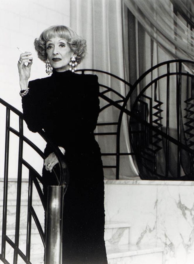 📷 Greg Gorman
Black & white photograph of Bette Davis standing on a white marble staircase leaning on a banister rail, the whole thing snazzy with its geometric black railing, wearing a black dress, ears bejeweled, cigarette in hand looking down towards the camera. She occupies the left hand side of the picture. On the right we see more of the marble, more of the banister railing and somewhat transparent grey curtains providing more geometric lines in their shadows and folds.