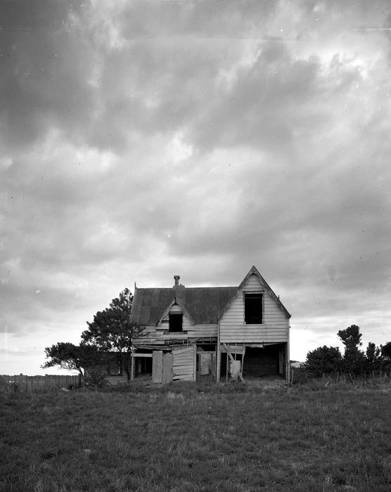 📷 Max Oettli 
Black & white photograph of a derelict house, still with its roof, apparently in the middle of nowhere. The house is positioned in the photograph just below the centre and a touch under the meeting point of grassland and sky, with shrubs and trees in a line to either side. But the sky is also a foreboding thing, cloudy, greys and white as if applied with brush strokes.